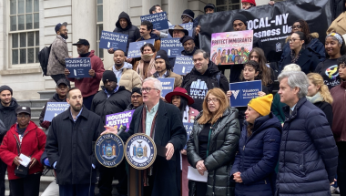 Alt text: Senator Brian Kavanagh speaks at a podium outside New York City Hall during a rally marking the launch of the Housing Access Voucher Program (HAVP). Standing with him are Assemblymember Linda Rosenthal, Assemblymember Jessica González-Rojas, Manhattan Borough President Brad Hoylman-Sigal, and Christopher Mann of Win. Housing advocates and community members stand on the steps behind them holding signs reading “Housing Is a Human Right” and banners from organizations including VOCAL-NY and Volunteer