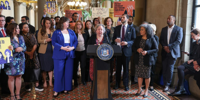 Senator Krueger stands at the podium with a group of legislators
