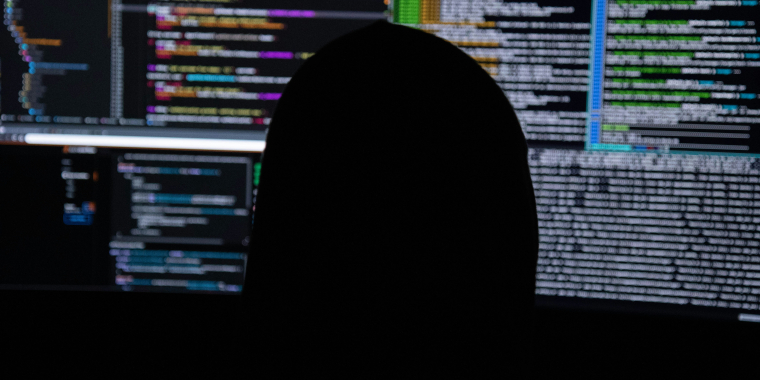 Photo image of a silhouette of a person sitting in front of multiple computer screens.