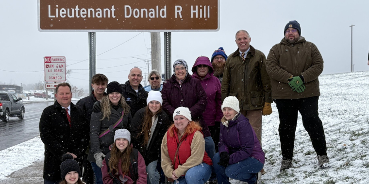 Group of people in front of brown ceremonial NYSDOT signs denoting "Lieutenant Donald R Hill Memorial Highway"