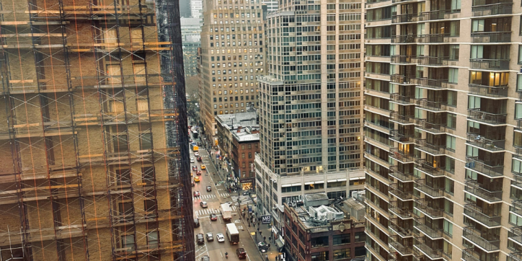A high-angle view looking down a New York City street canyon on an overcast, foggy day. On the left, a historic brick building is covered in construction scaffolding. On the right, a modern high-rise residential building with balconies. The street below is active with yellow taxis, pedestrians, and a Chick-fil-A visible at street level. Skyscrapers disappear into the fog in the background.